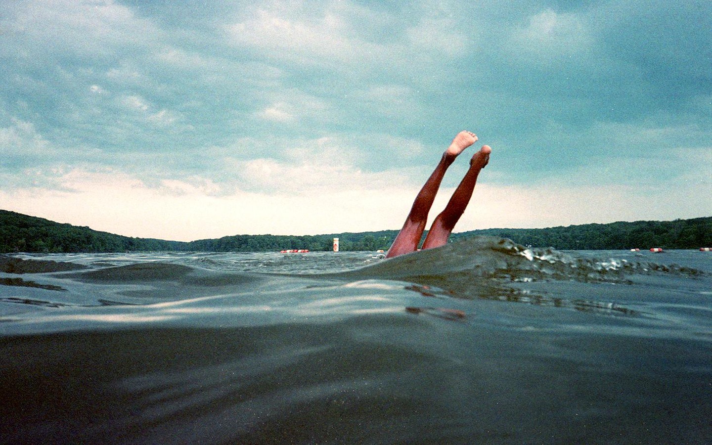 Two legs are visible sticking out of the water in a lake with a forested shore under a cloudy sky.