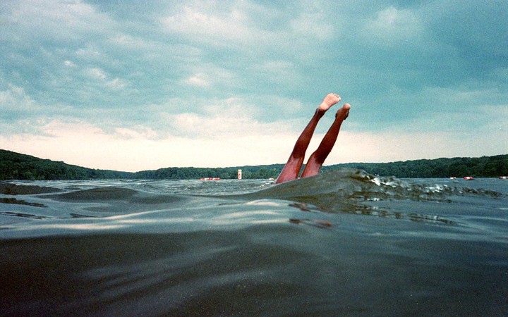 Two legs are visible sticking out of the water in a lake with a forested shore under a cloudy sky.