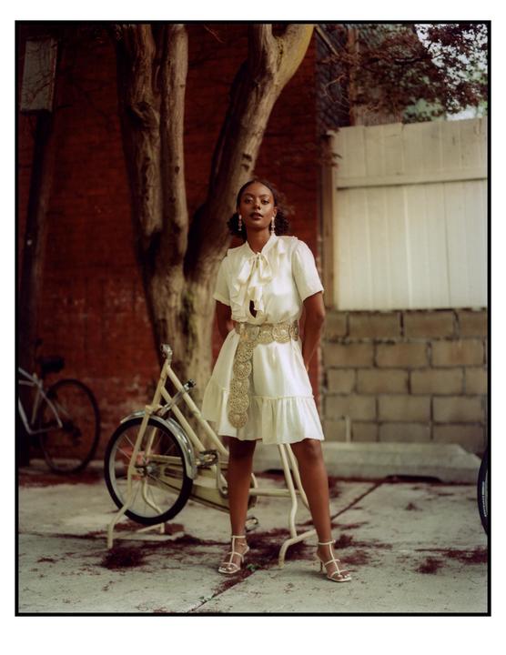 A woman in a white dress and gold belt stands in front of a tree and a bicycle.