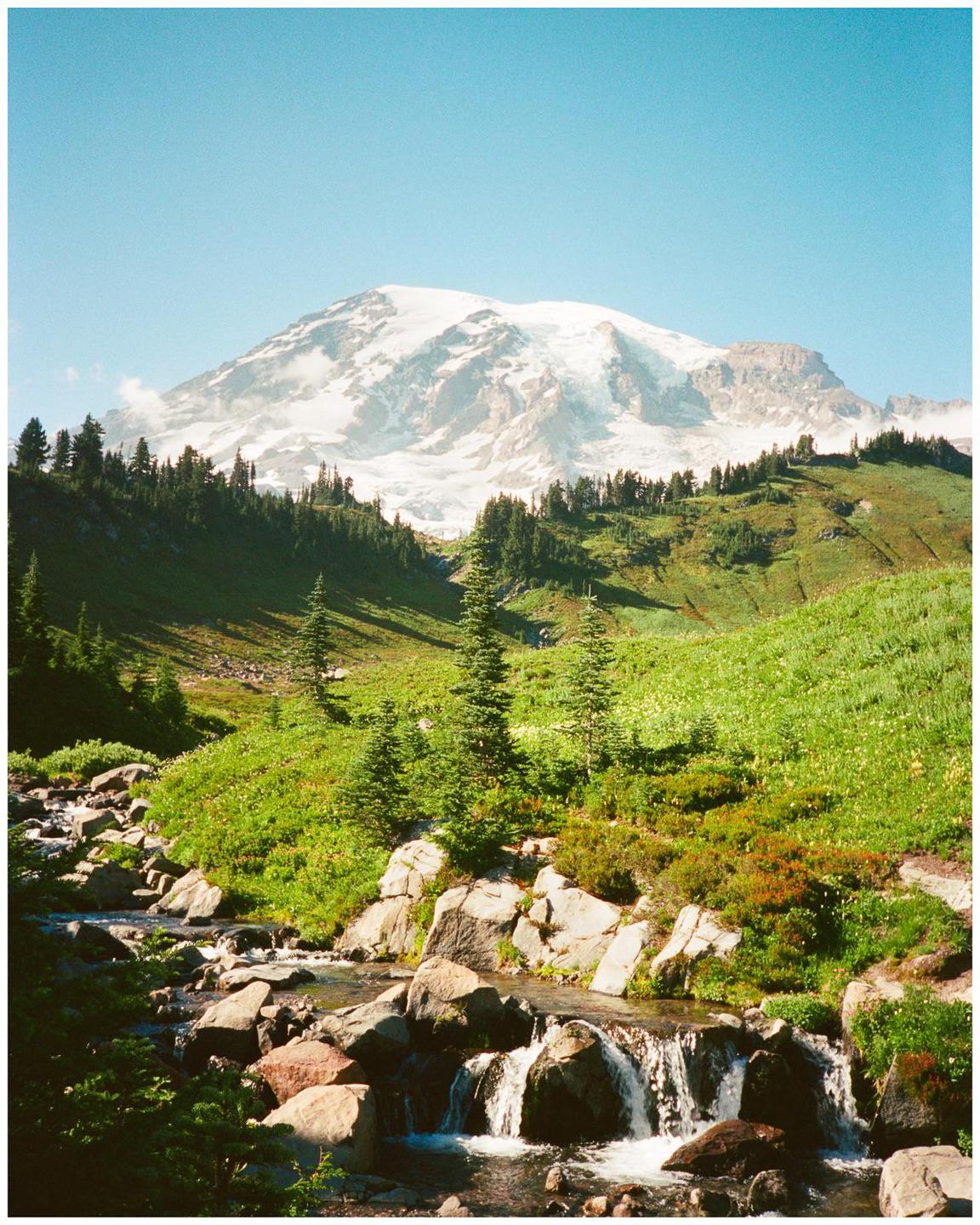 A snow-capped mountain rises above a green valley with a rocky stream and evergreen trees.