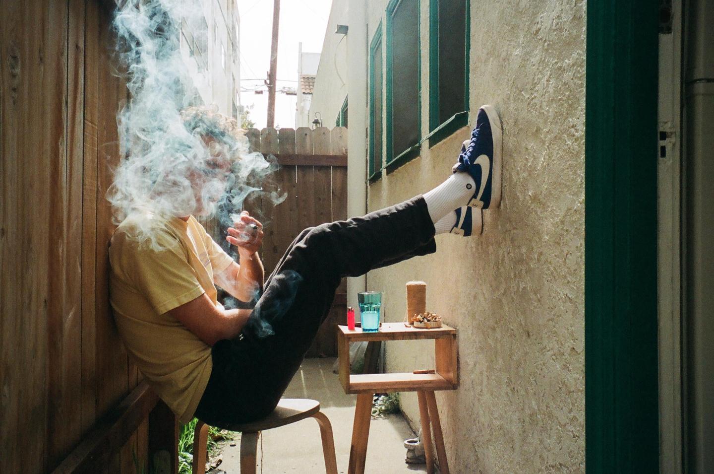 A man sits on a stool with his feet up against a wall, smoking a cigarette with a lot of smoke around his head.