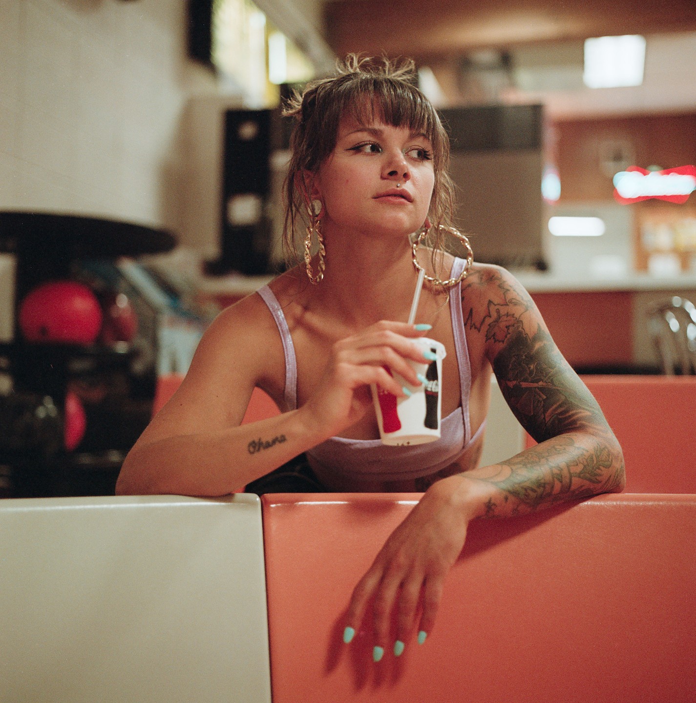 A woman with tattoos on her arms sits in a booth, holding a drink with a straw.