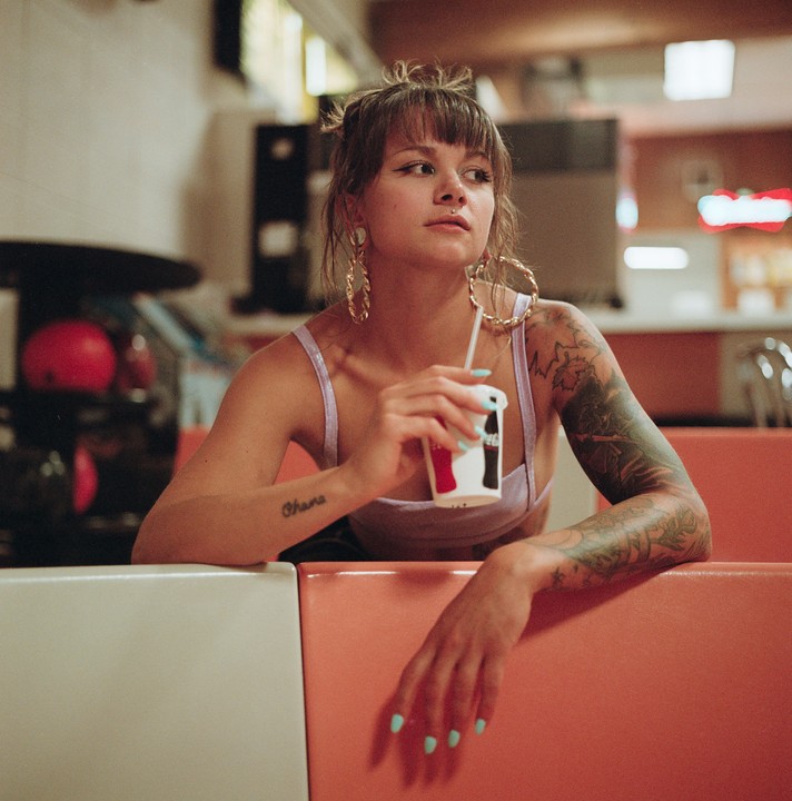A woman with tattoos on her arms sits in a booth, holding a drink with a straw.