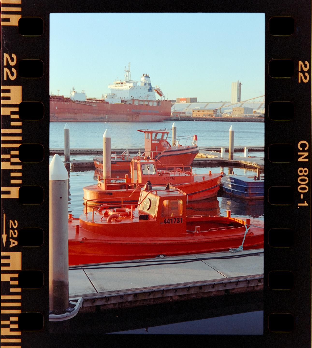 Several small red boats are docked in a harbor with a large cargo ship in the background.