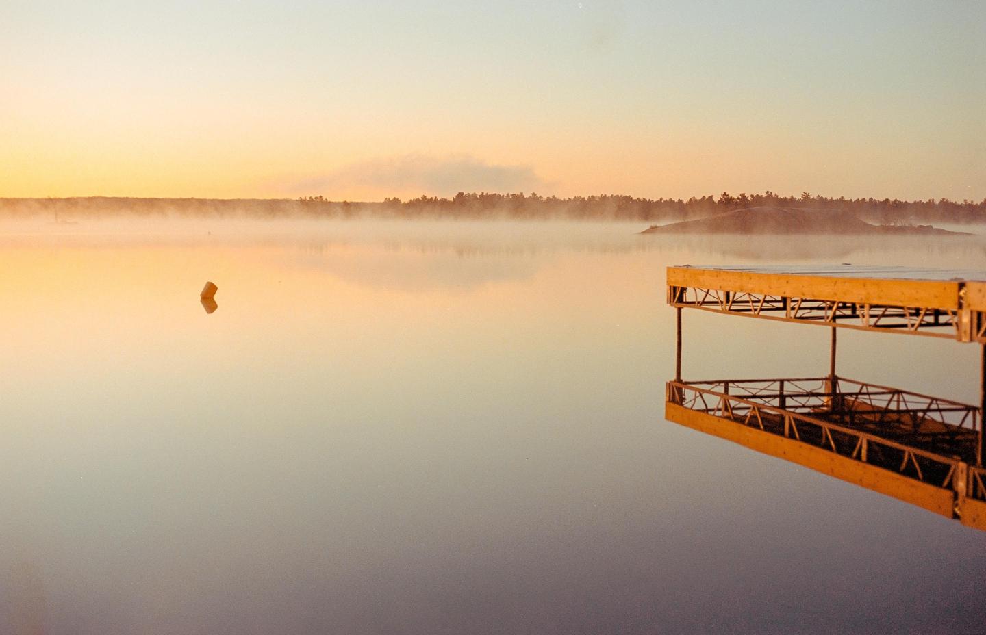 A wooden dock extends into a misty lake at sunrise, with a buoy floating in the distance and a forested shoreline visible through the fog.