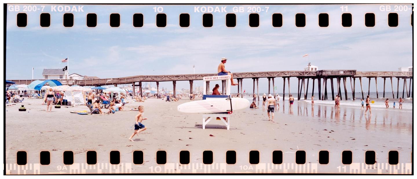A lifeguard sits on a high chair on a crowded beach with a pier in the background.