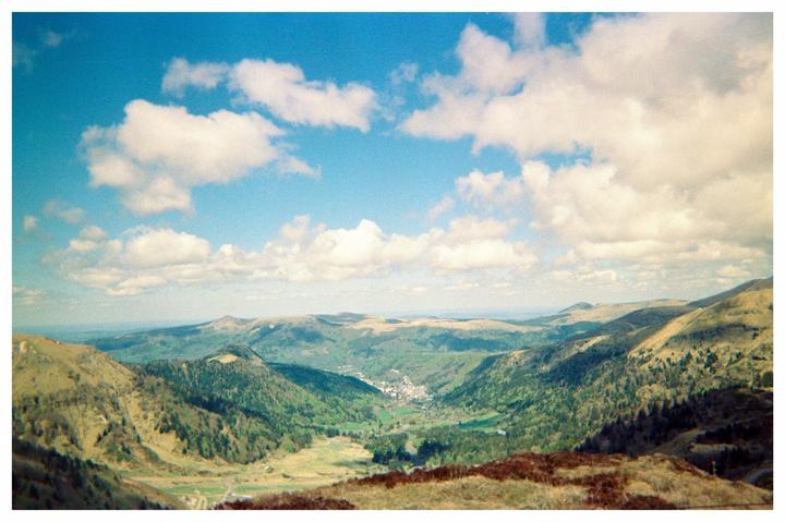 A small village is nestled in a valley surrounded by rolling green hills under a blue sky with white clouds.