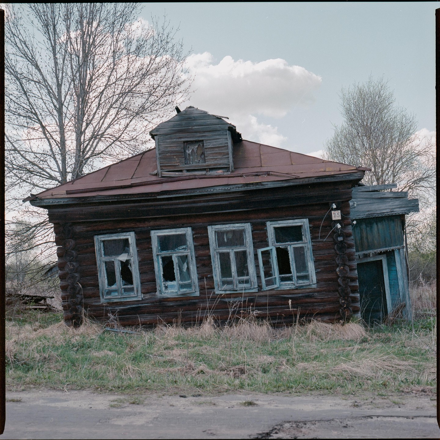 An old wooden house with broken windows and a rusty roof stands next to a dirt road.