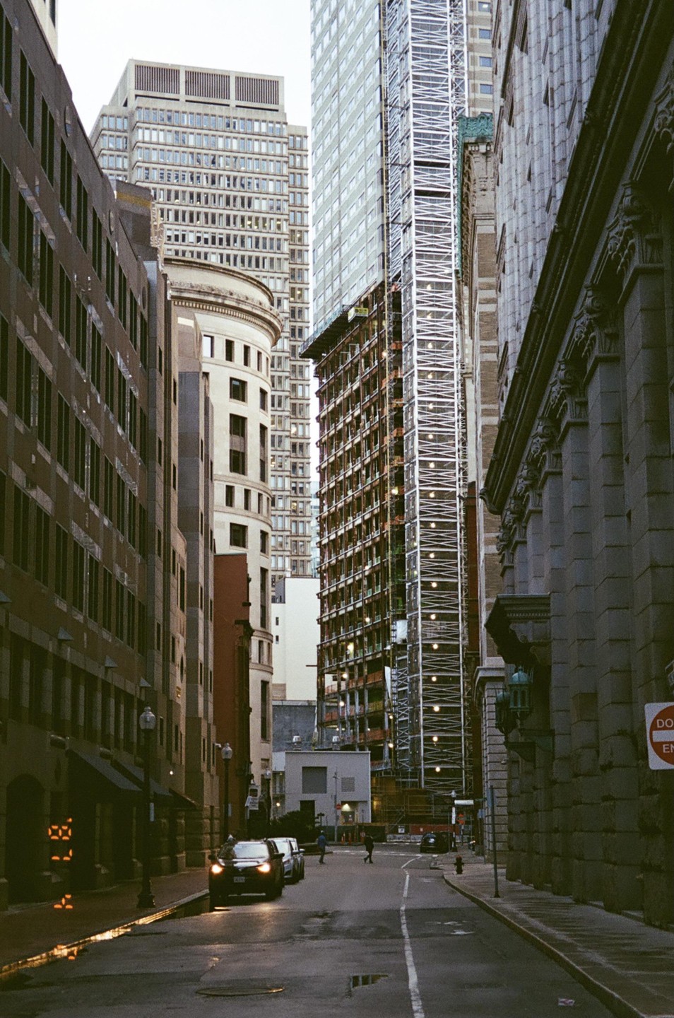 A narrow street is lined with tall buildings, some under construction, with a few cars and pedestrians visible.