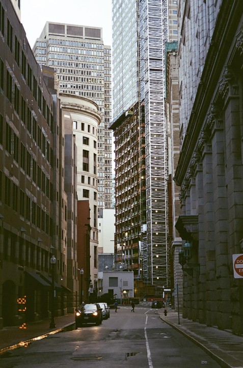 A narrow street is lined with tall buildings, some under construction, with a few cars and pedestrians visible.