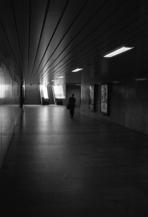 A person walks away down a long, dark underground passage towards a bright light and stairs.