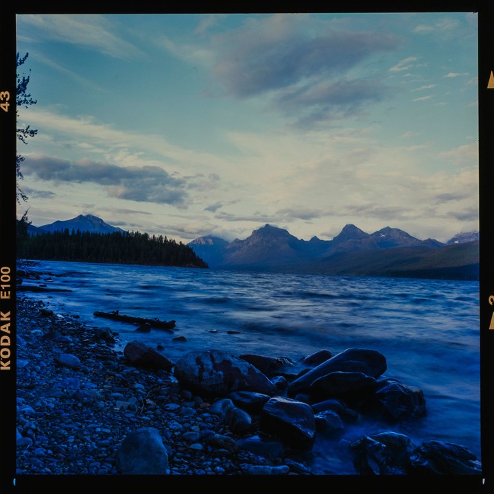 A lake with mountains and a forest in the background is viewed from a rocky shore under a cloudy sky.