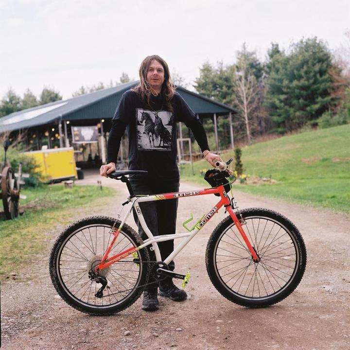 A man with long hair stands on a dirt path next to a red and white bicycle.