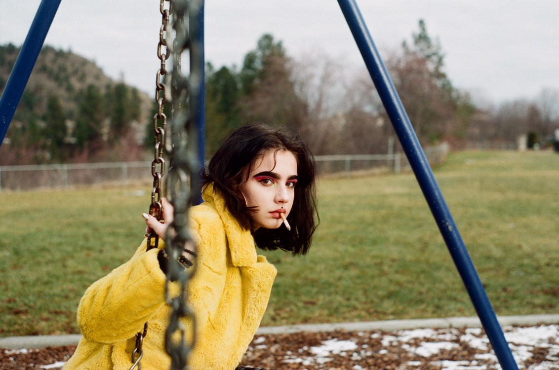 A woman in a yellow coat sits on a swing set with a cigarette in her mouth, looking towards the viewer.