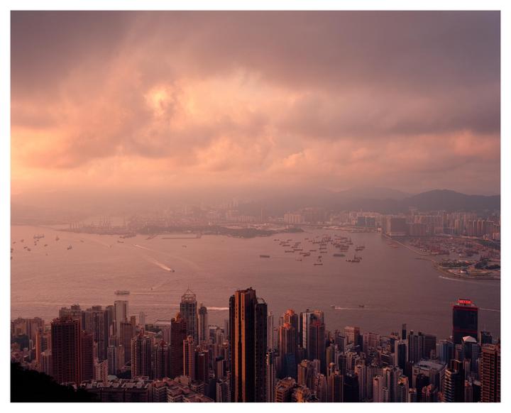 A panoramic view of a city with many buildings and a body of water with boats under a cloudy, colorful sky.