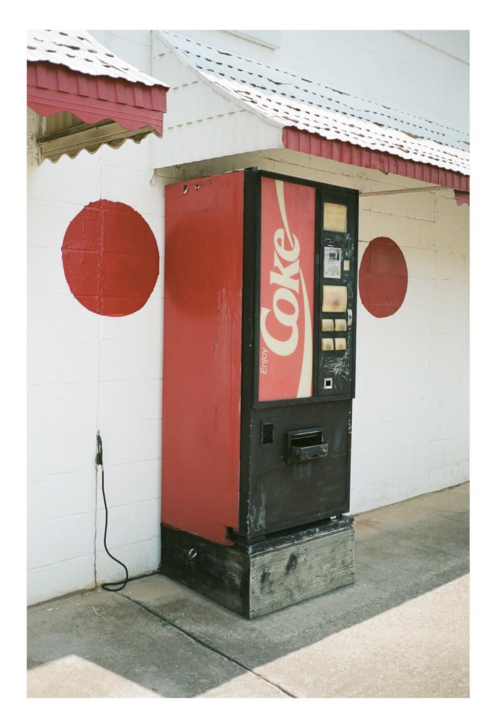 A red Coca-Cola vending machine stands against a white wall with two red circles painted on it.