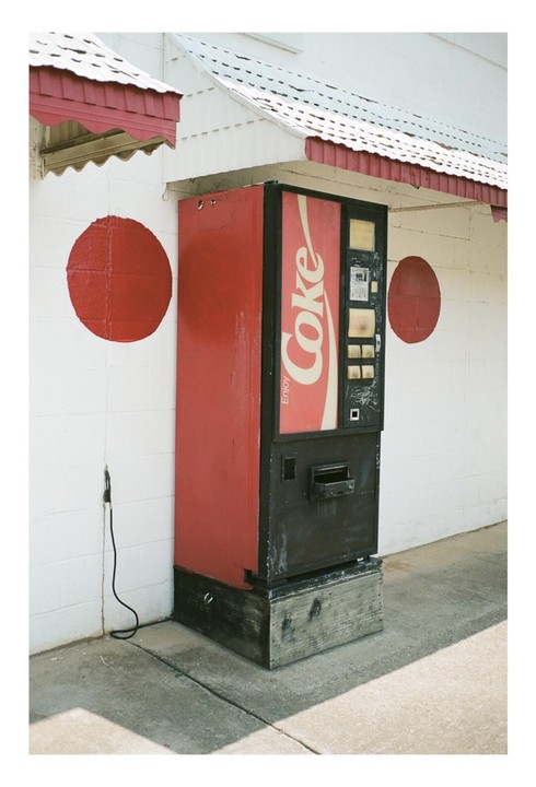A red Coca-Cola vending machine stands against a white wall with two red circles painted on it.