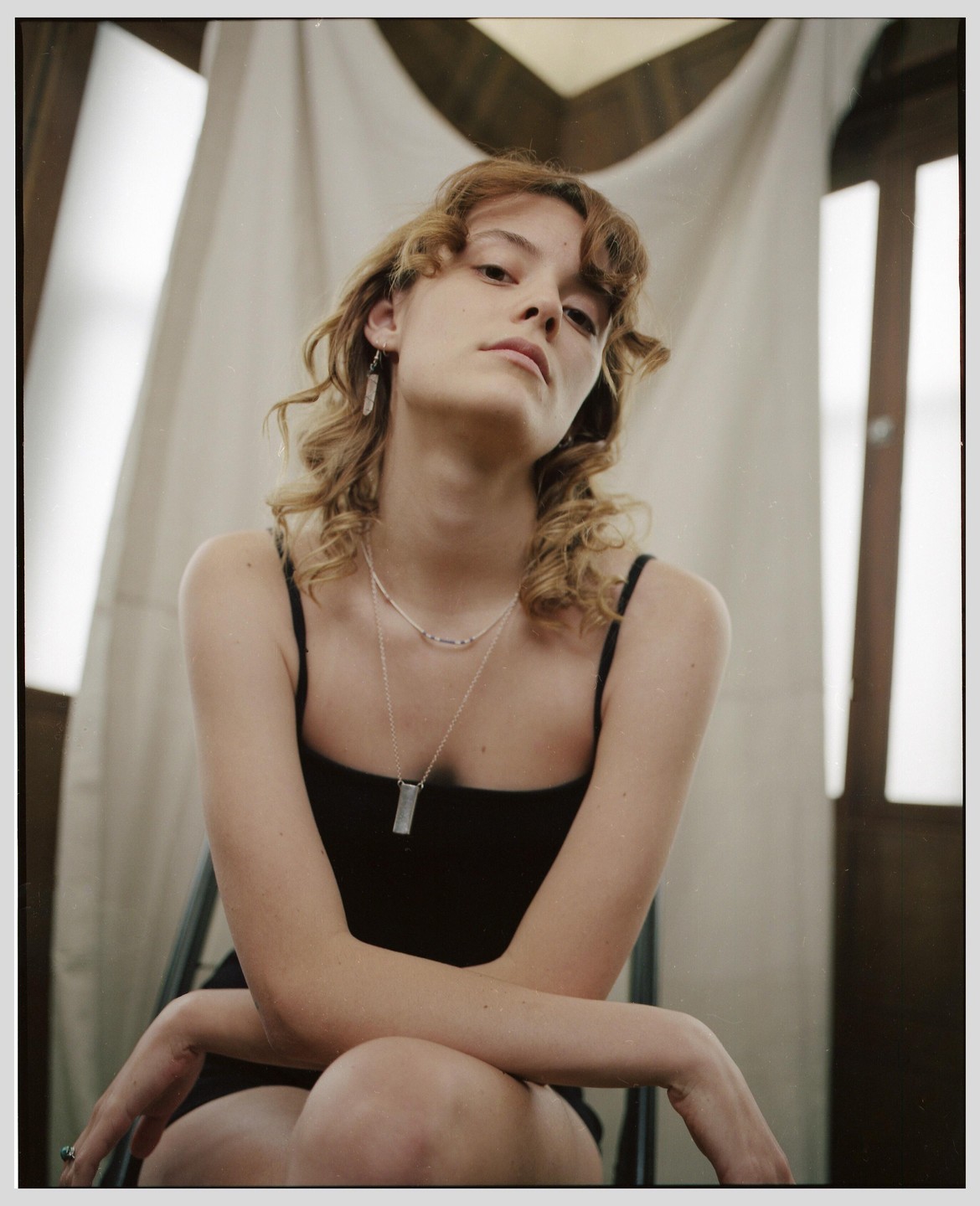 A woman with curly hair wearing a black top and necklaces sits with her arms crossed, looking directly at the viewer.