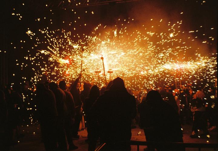 A crowd of people watches a correfoc with sparks flying through the night sky.