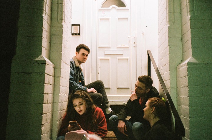 Four young adults are sitting on steps in an entryway, with one woman looking at her phone and two men smoking.