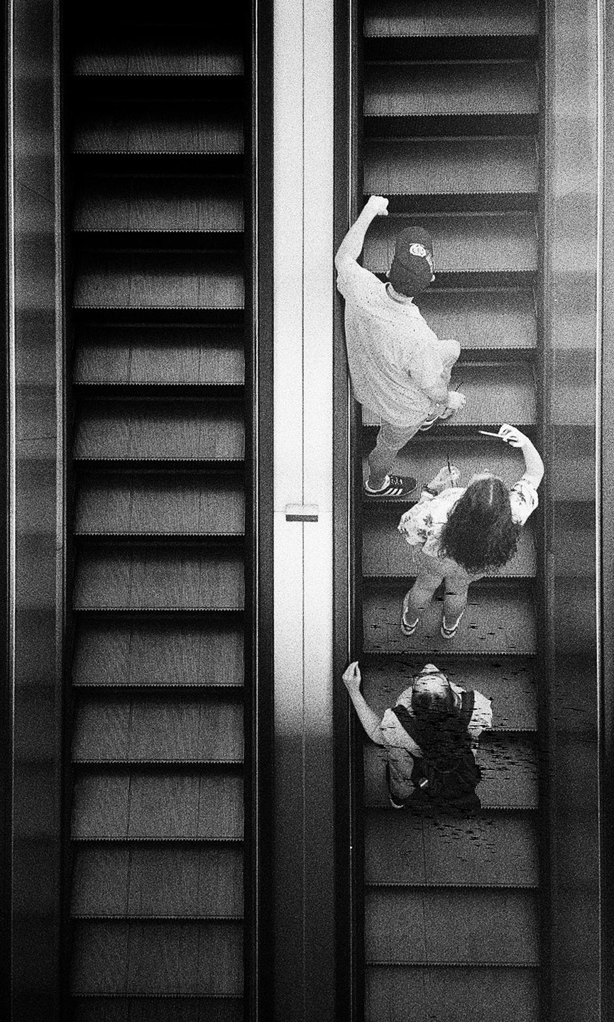 Three people are riding an escalator next to an empty escalator, viewed from above.