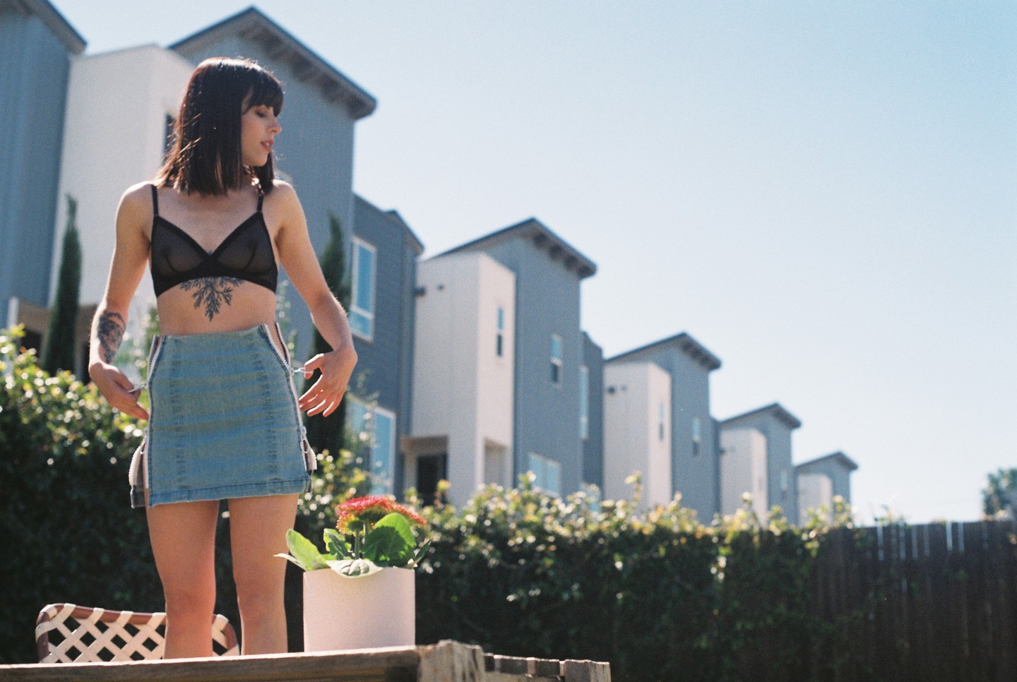A woman in a bra and denim skirt stands in a backyard with townhouses in the background.