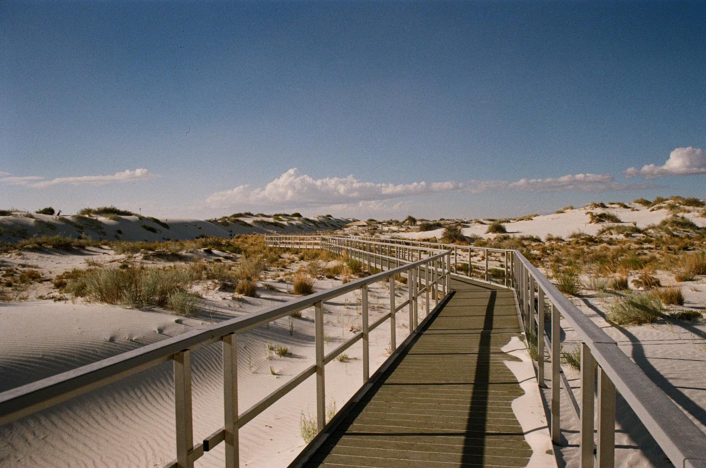 A long boardwalk with railings winds through white sand dunes covered in sparse vegetation under a blue sky with scattered clouds.