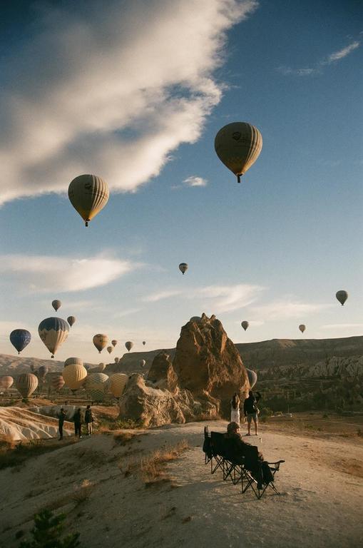 Hot air balloons float in a blue sky above a rocky landscape with people watching from a hill.
