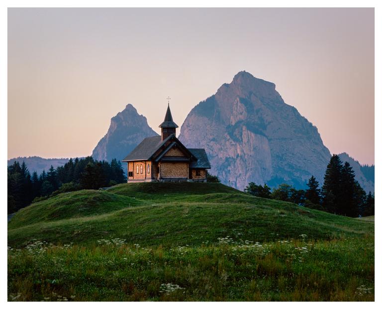 A small wooden church with a steeple stands on a grassy hill with large mountains in the background.