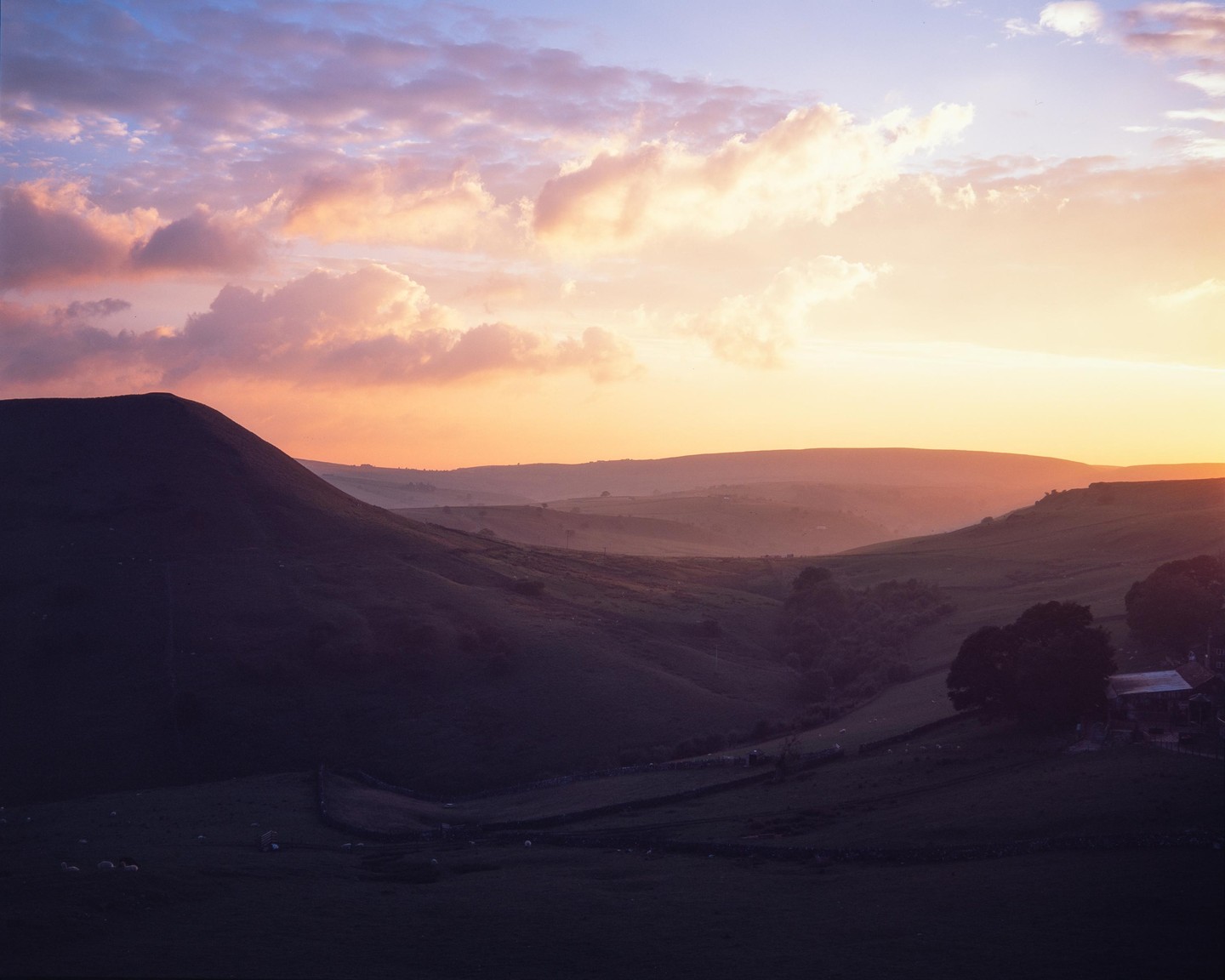 A dramatic sunset casts a warm glow over rolling hills and a valley with a farm.