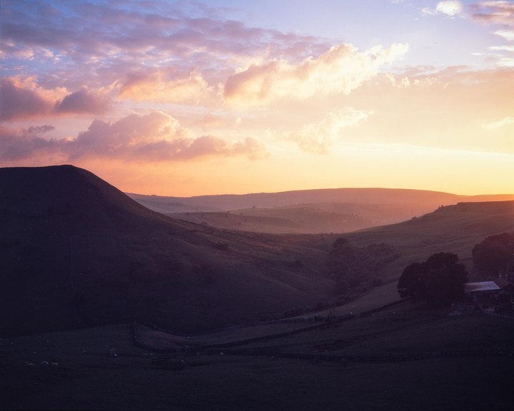 A dramatic sunset casts a warm glow over rolling hills and a valley with a farm.