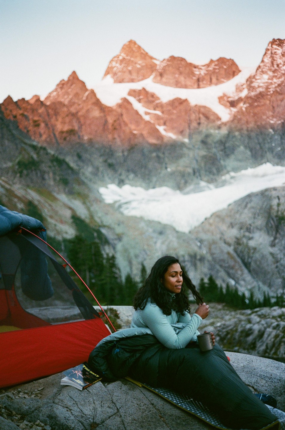 A woman sits in a sleeping bag next to a tent with snow-capped mountains in the background.