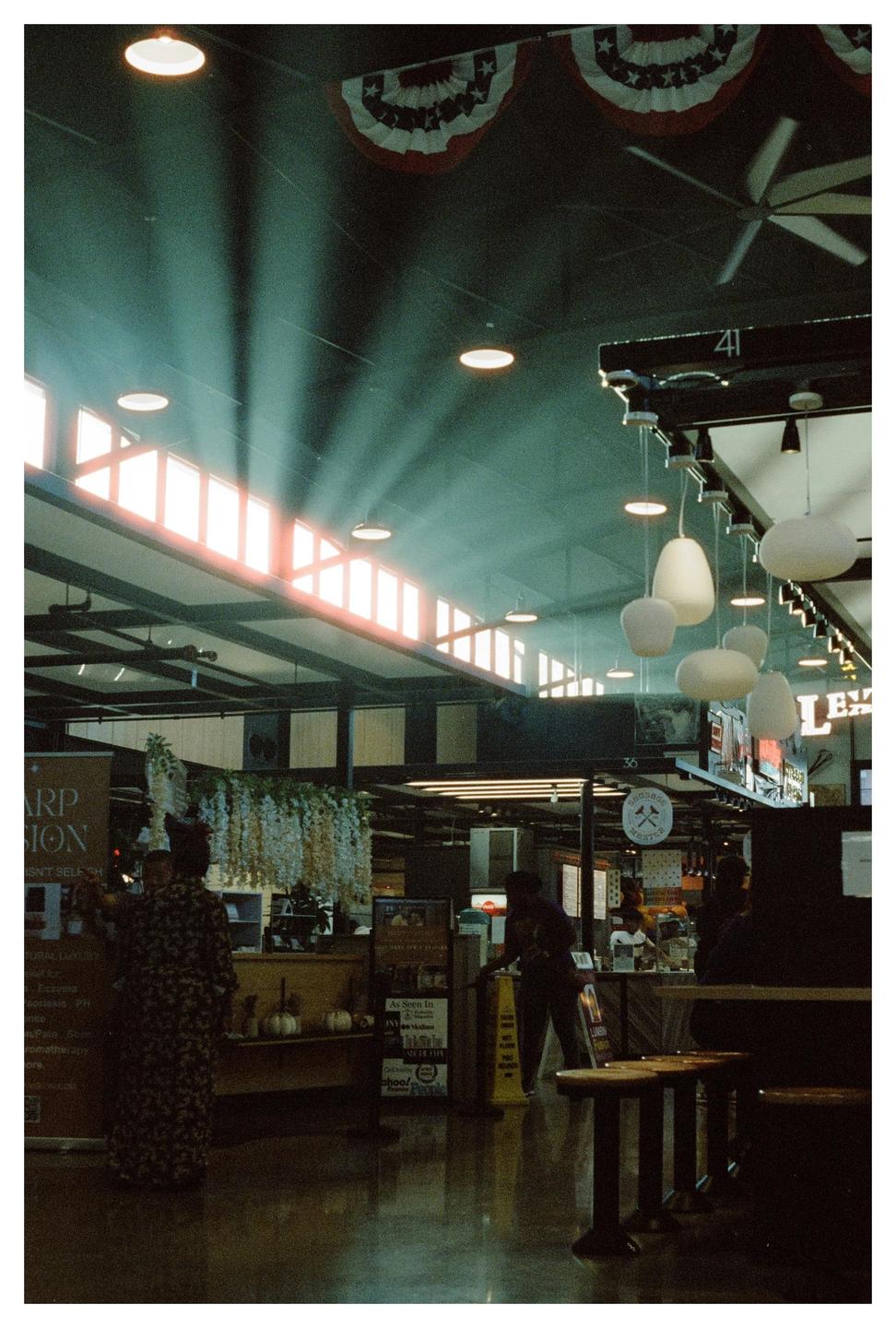Sunlight streams through high windows into a large indoor market with people, stalls, and hanging decorations.