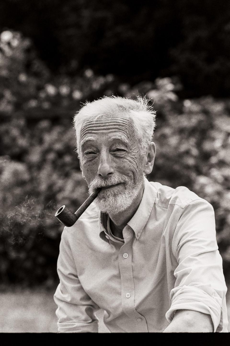 A man with a white beard and hair smiles while smoking a pipe.