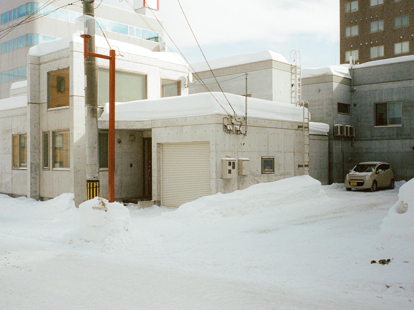 A small car is parked next to a snow-covered building with a utility pole in the foreground.