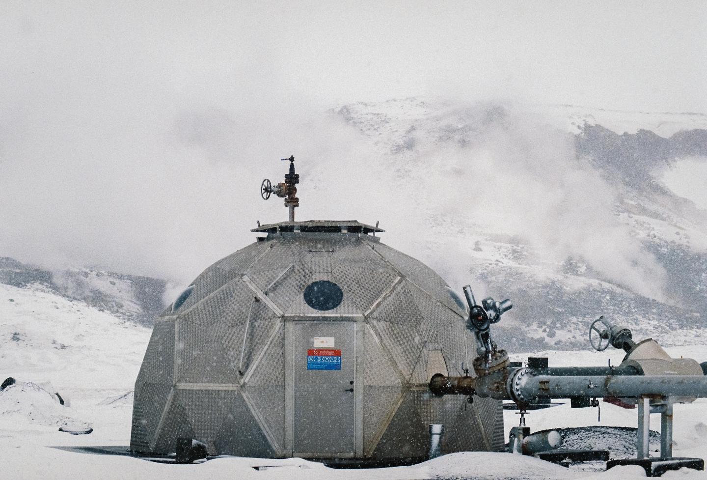 A geodesic dome structure with pipes attached to its side stands in a snowy, mountainous landscape.