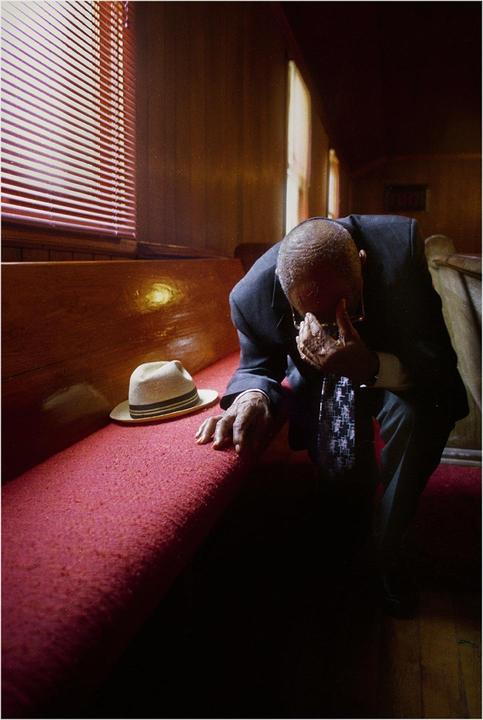 A man in a suit sits with his head in his hand next to a hat on a red pew.
