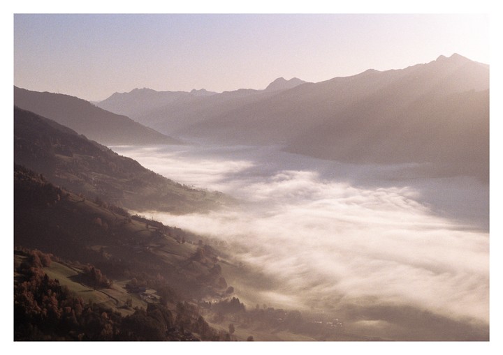 A valley filled with fog is surrounded by mountains and hills with autumn trees.