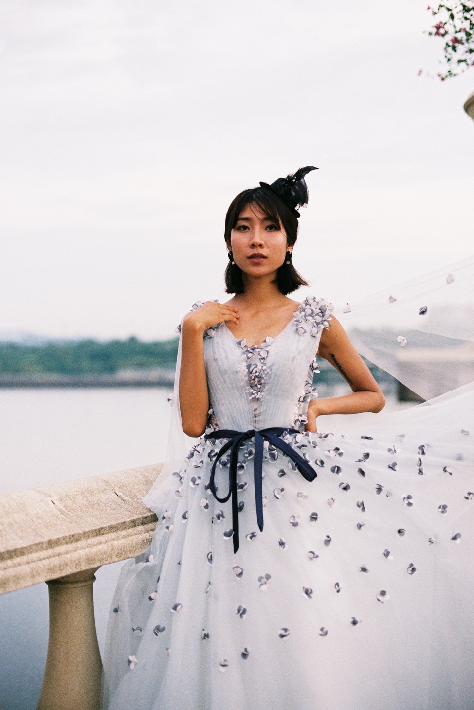 A woman in a light blue gown with floral appliques and a dark blue ribbon stands on a stone balcony overlooking water.