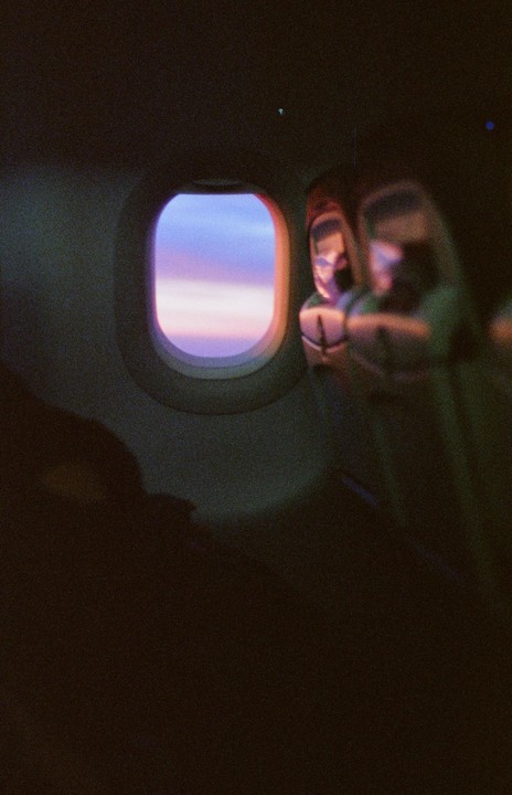 The view from an airplane window shows a colorful sunset sky and the reflection of a passenger.