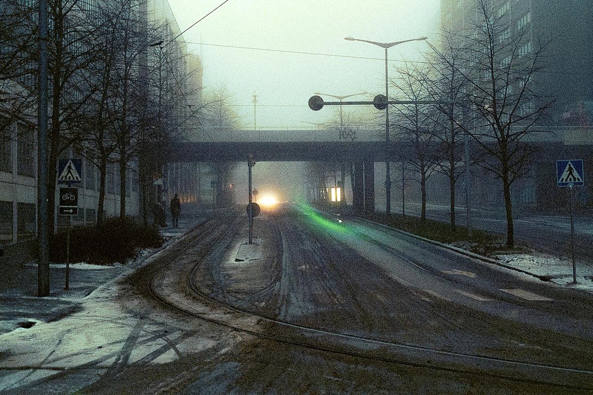 A street with tram tracks and bare trees is covered in snow and fog, with a bright light visible in the distance under a bridge.