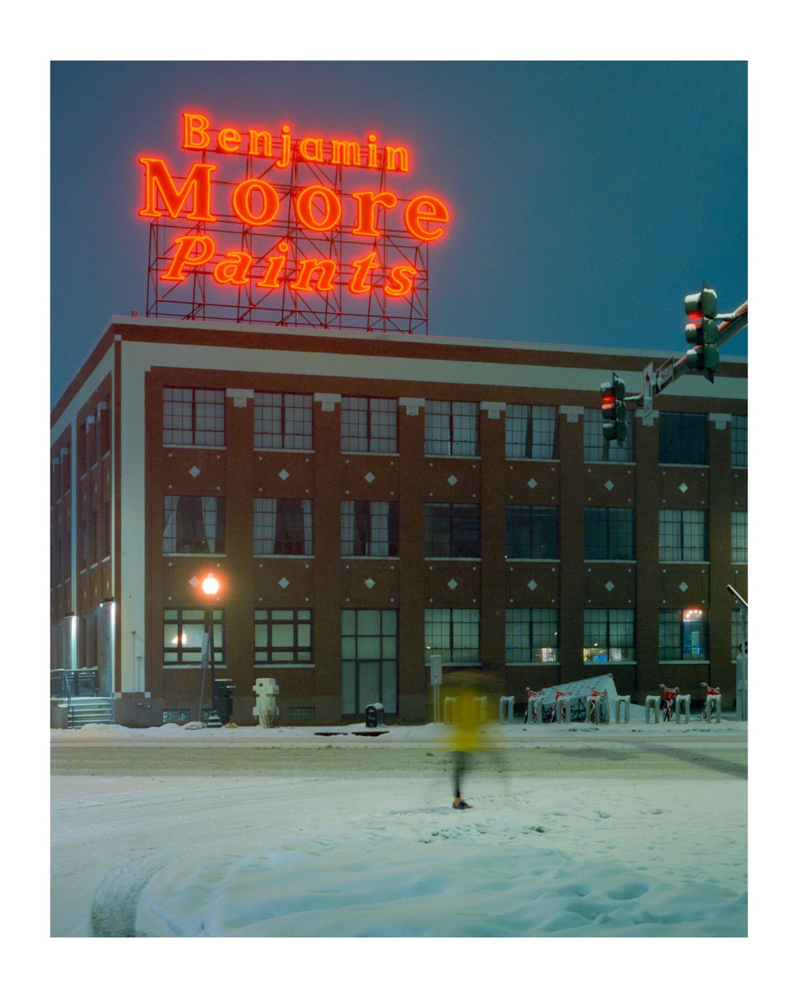 A blurry person in a yellow jacket walks across a snow-covered street at night, with a large neon sign for Benjamin Moore Paints on top of a brick building in the background.