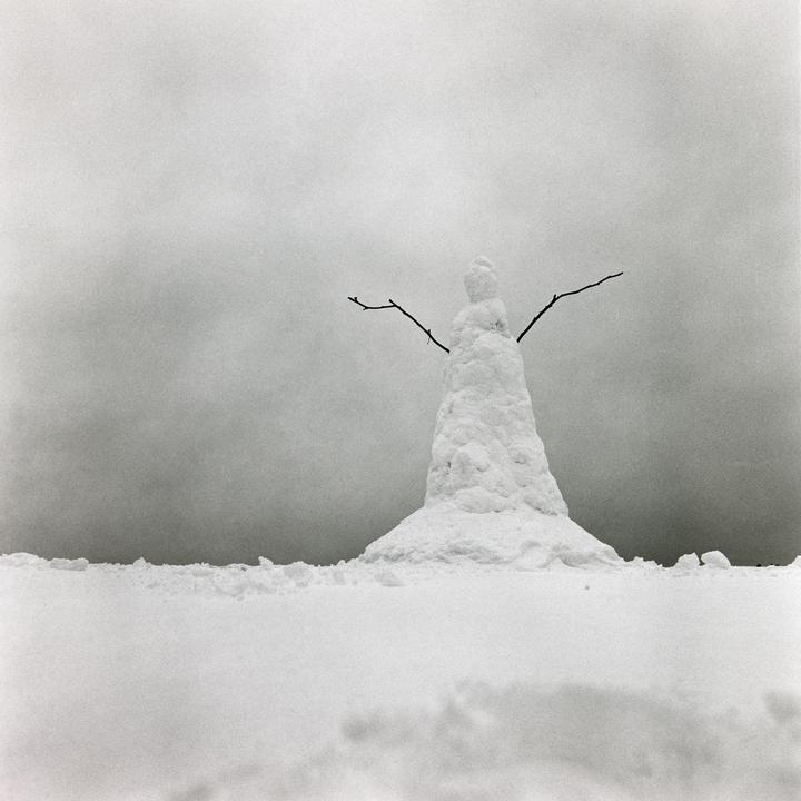 A snowman with stick arms stands in a field of snow under a cloudy sky.