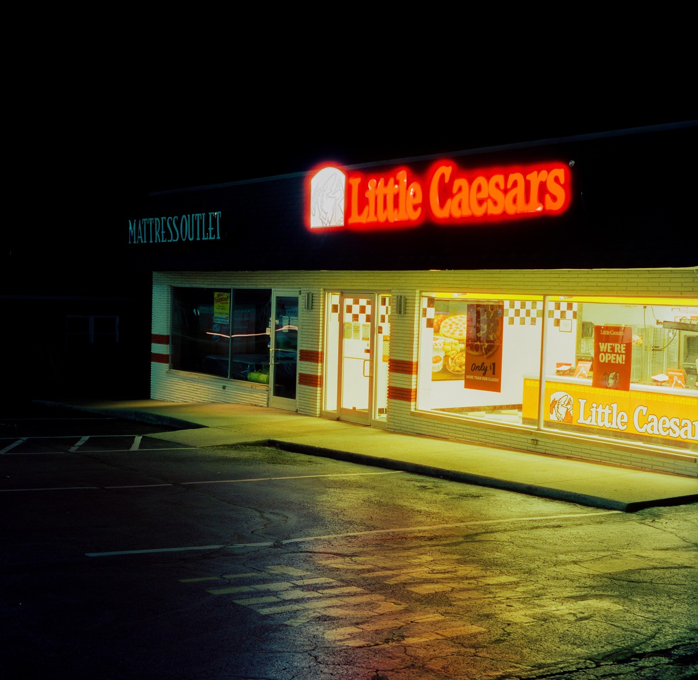 A Little Caesars pizza restaurant with a neon sign is illuminated at night next to a Mattress Outlet.