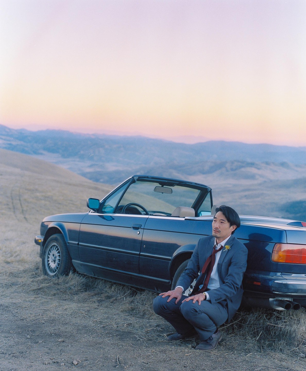 A man in a suit crouches next to a convertible car on a hill overlooking a valley at sunset.