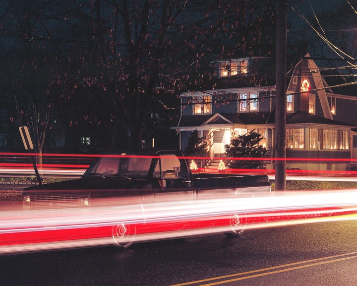 A black pickup truck is parked on a street at night with light trails from passing vehicles and a house decorated with lights in the background.