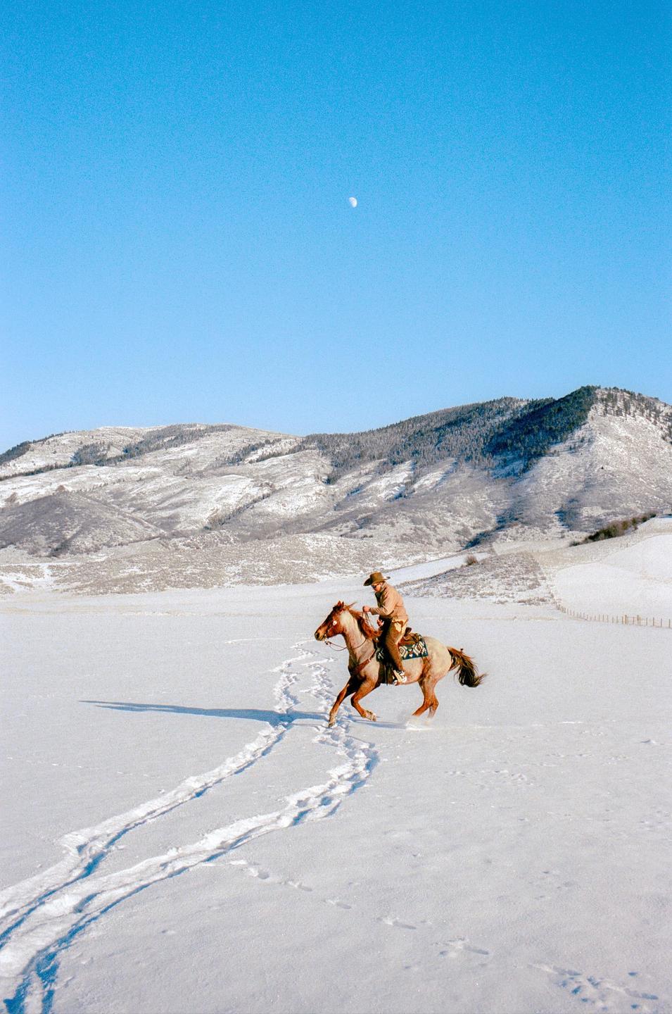 A man on horseback rides through a snow-covered field with mountains in the background under a clear blue sky with a visible moon.