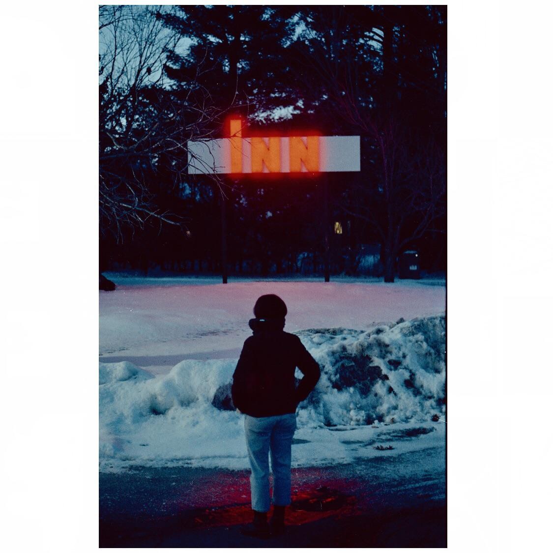 A person stands in the snow at dusk, looking at a neon sign that reads "INN."