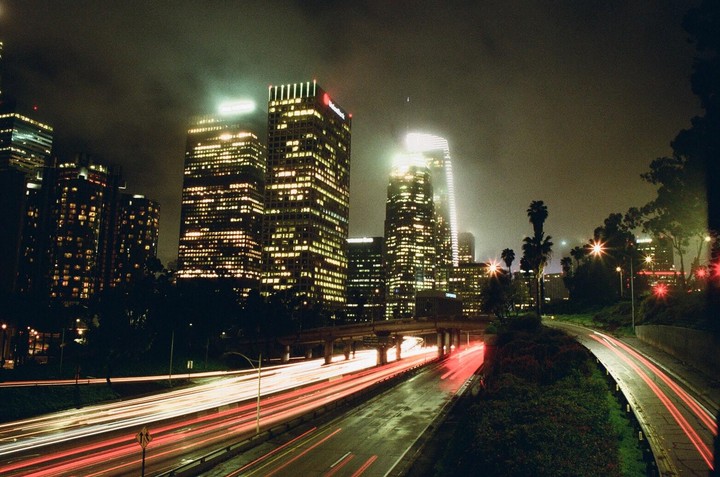 A highway with light trails from cars passes by illuminated skyscrapers at night.