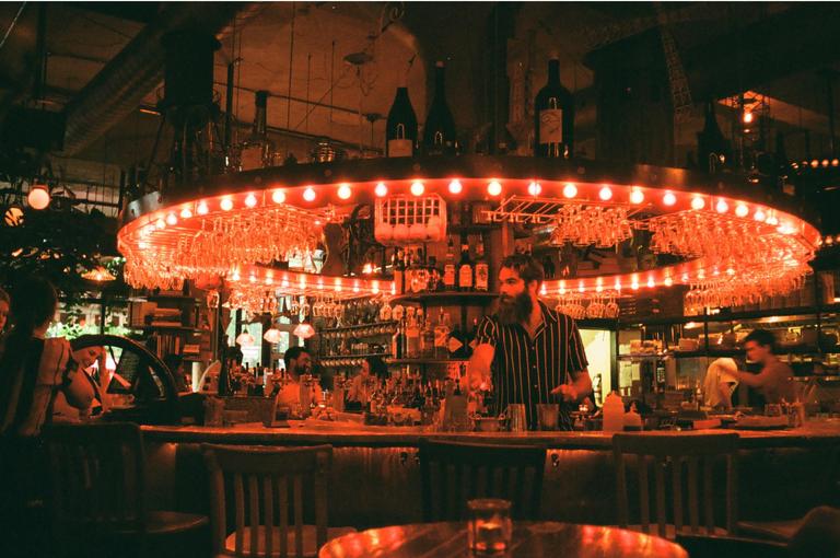 A bearded man works behind a bar with a circular light fixture above it.